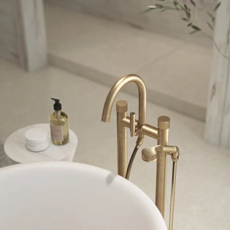 Modern gold freestanding bathtub faucet and white tub with bath products on a side table in a minimalist bathroom.