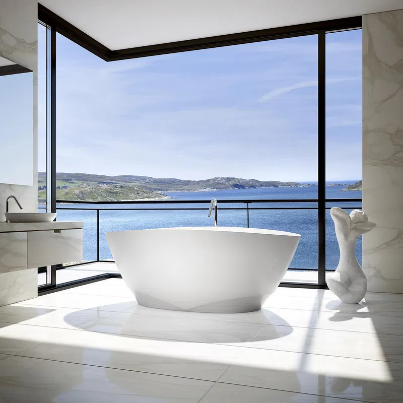Modern bathroom with freestanding white bathtub and large windows overlooking ocean and hills.