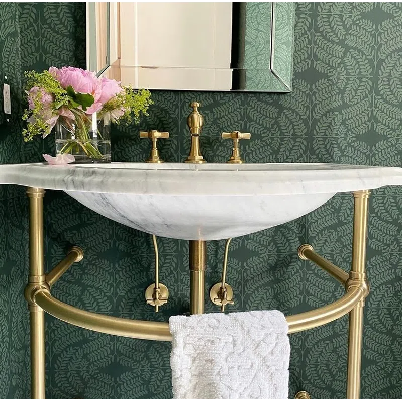 Elegant bathroom sink with brass fixtures, marble countertop, green leaf-patterned wallpaper, and pink flowers in vase.