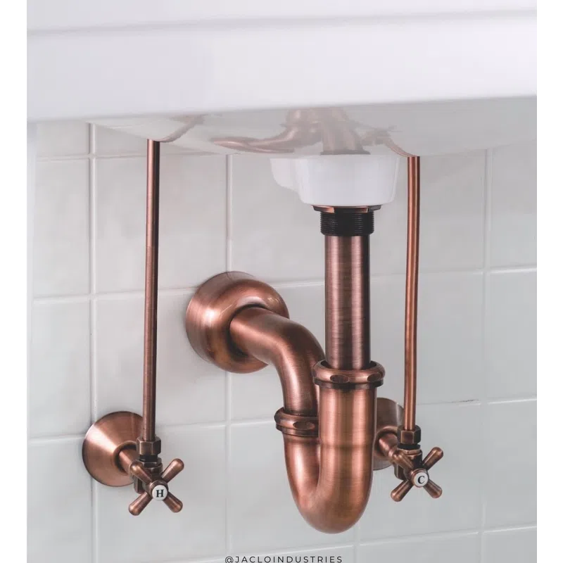 Copper plumbing pipes and fittings under a white sink with hot and cold water valves against tiled wall.