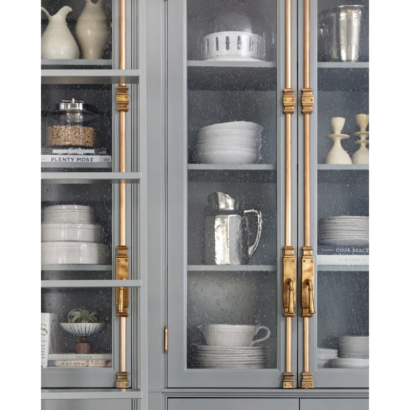 Gray kitchen cabinet with textured glass doors displaying white dishes, pitchers, and gold hardware handles.