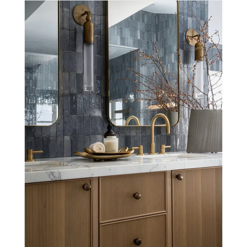 Modern bathroom vanity with brass fixtures, marble countertop, dark tiled backsplash, and decorative vase with branches.