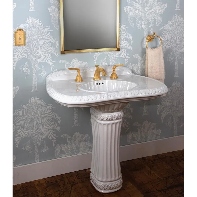 Elegant white pedestal sink with gold fixtures against blue palm tree wallpaper and wooden floor.