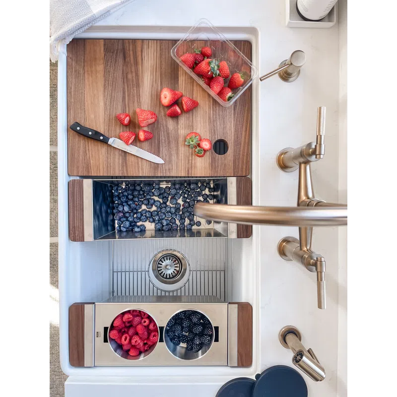 Wooden cutting board with sliced strawberries, bins of blueberries, raspberries, and blackberries in a white kitchen sink with brass faucet.