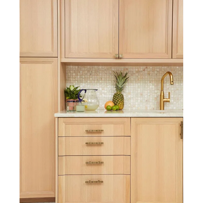 Modern kitchen with light wood cabinets, mosaic tile backsplash, gold faucet, and fresh fruit on white countertop.