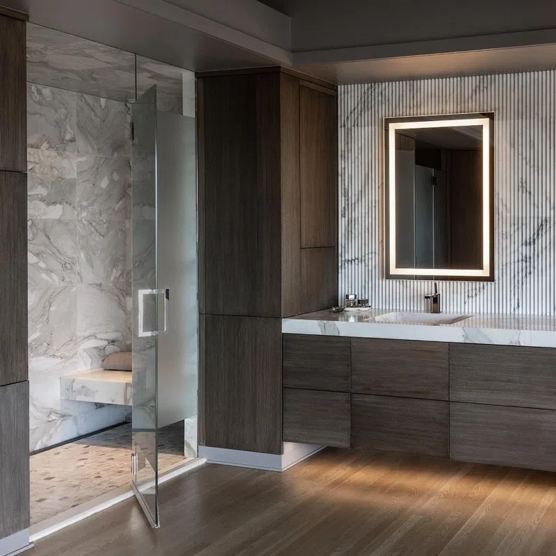 Modern bathroom with wooden cabinets, marble countertops, illuminated mirror, and glass shower with marble tiles.