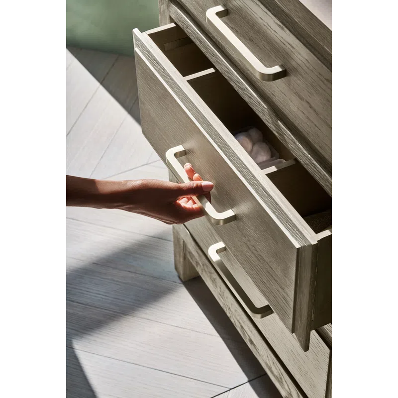 Hand opening a wooden drawer with beige handles showing interior storage and sunlight on wood floor.