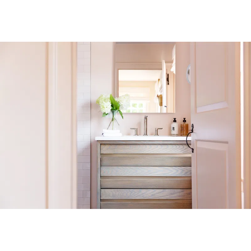 Light wood bathroom vanity with double faucets, mirror, white flowers in vase, and soap dispensers.