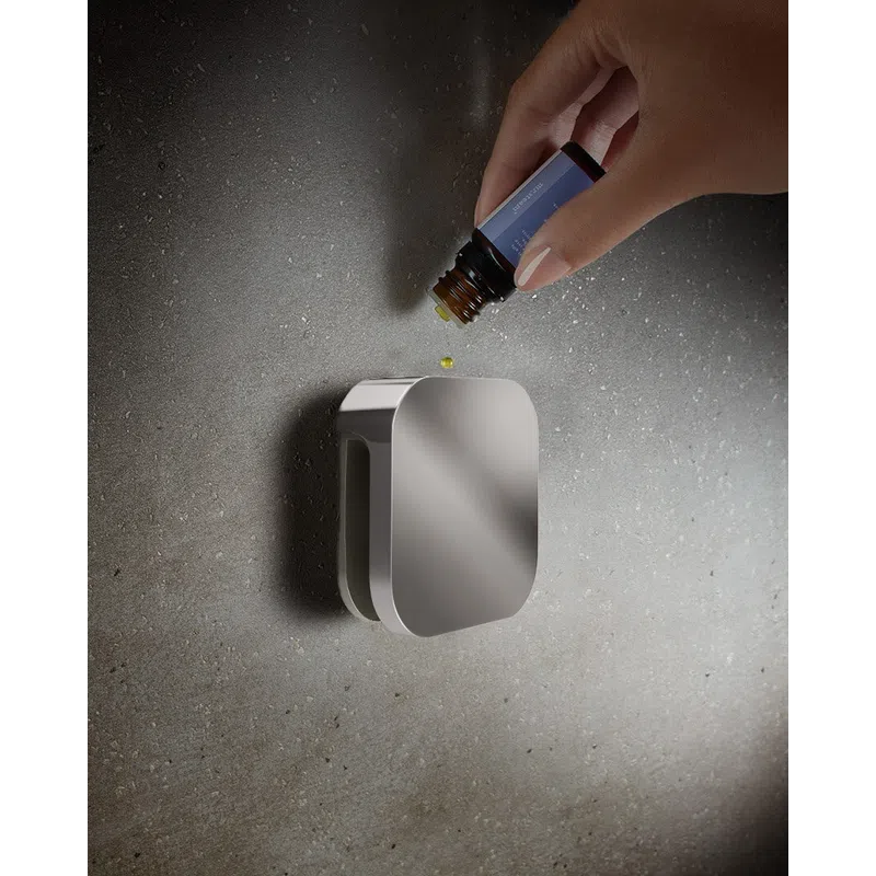 Hand drops essential oil from a brown bottle onto a sleek silver diffuser on a textured gray surface.
