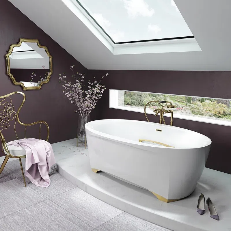 Modern bathroom with white freestanding tub, gold fixtures, skylight, decorative chair, and floral accents.
