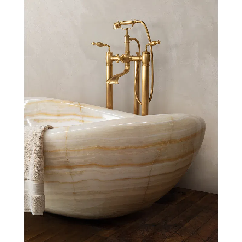 Close-up of a beige marble bathtub with a beige towel and vintage gold faucet on textured wall and wooden floor.