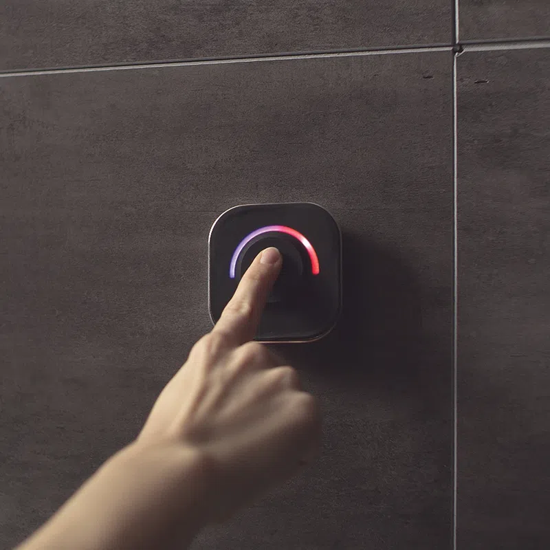 Hand adjusting a modern thermostat dial with blue and red temperature indicators on a dark tiled wall.