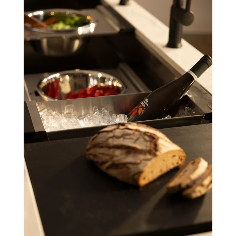 Loaf of crusty bread sliced on counter next to a wine bottle chilling in ice and bowls of fresh vegetables.