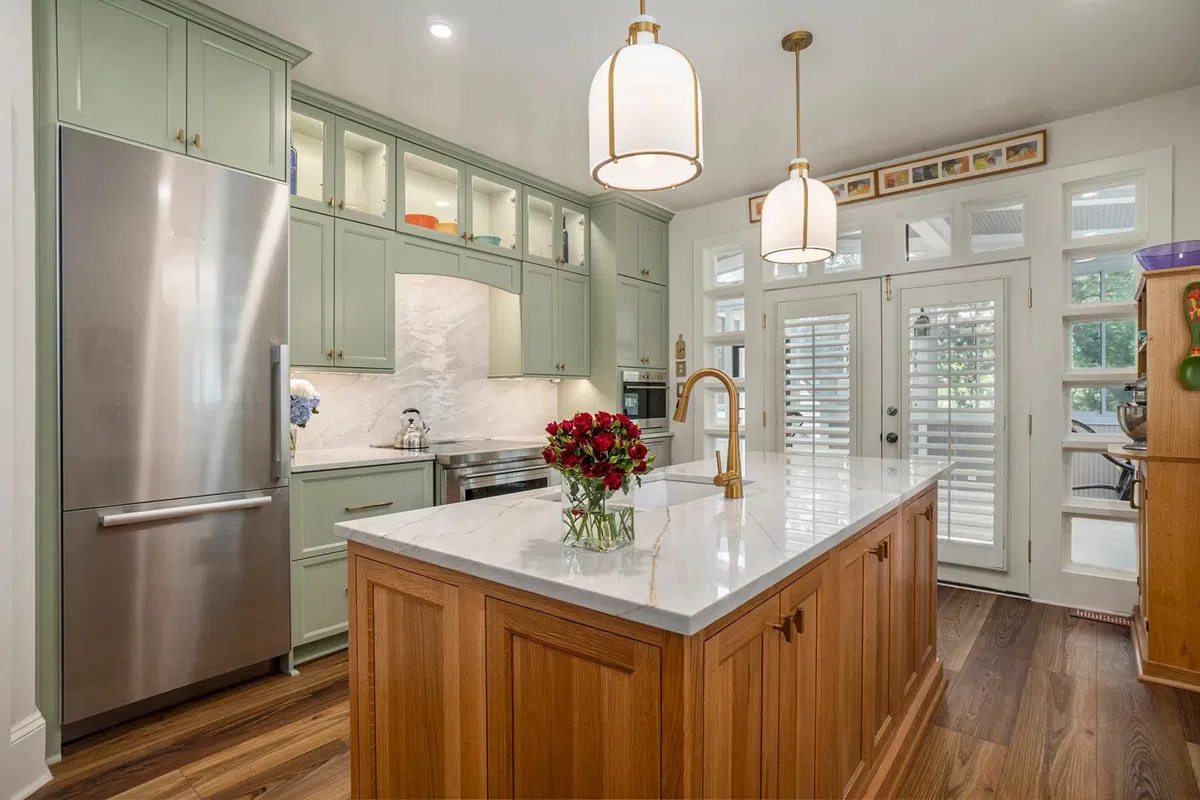 Modern kitchen with green cabinets, wooden island, marble countertops, pendant lights, and stainless steel refrigerator.