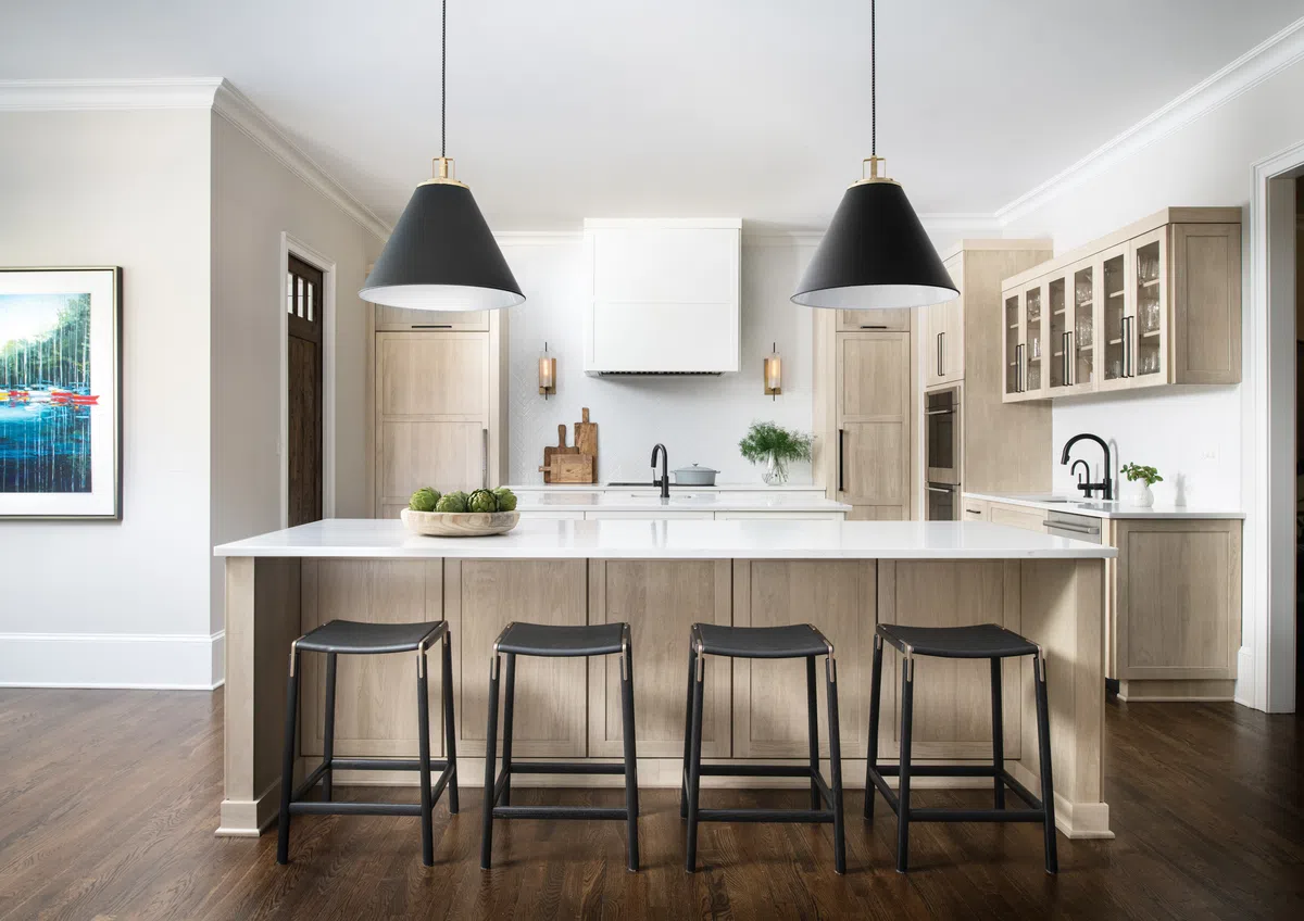 Modern kitchen with a large island, four black stools, pendant lights, and wood flooring under natural light.