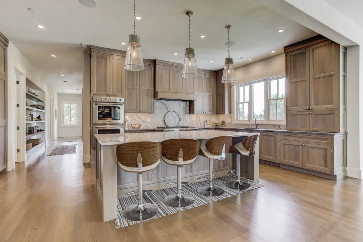 Modern kitchen with wood cabinetry, marble countertops, four bar stools, pendant lights, and hardwood floors.