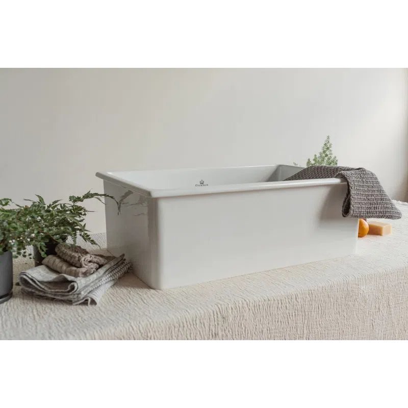 White ceramic farmhouse sink displayed on textured surface with towels, plant, soap, and gray cloth.