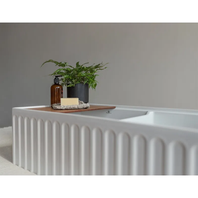 Modern white bathtub with textured sides, wooden tray holding soap, brown bottle, and a green potted fern plant.