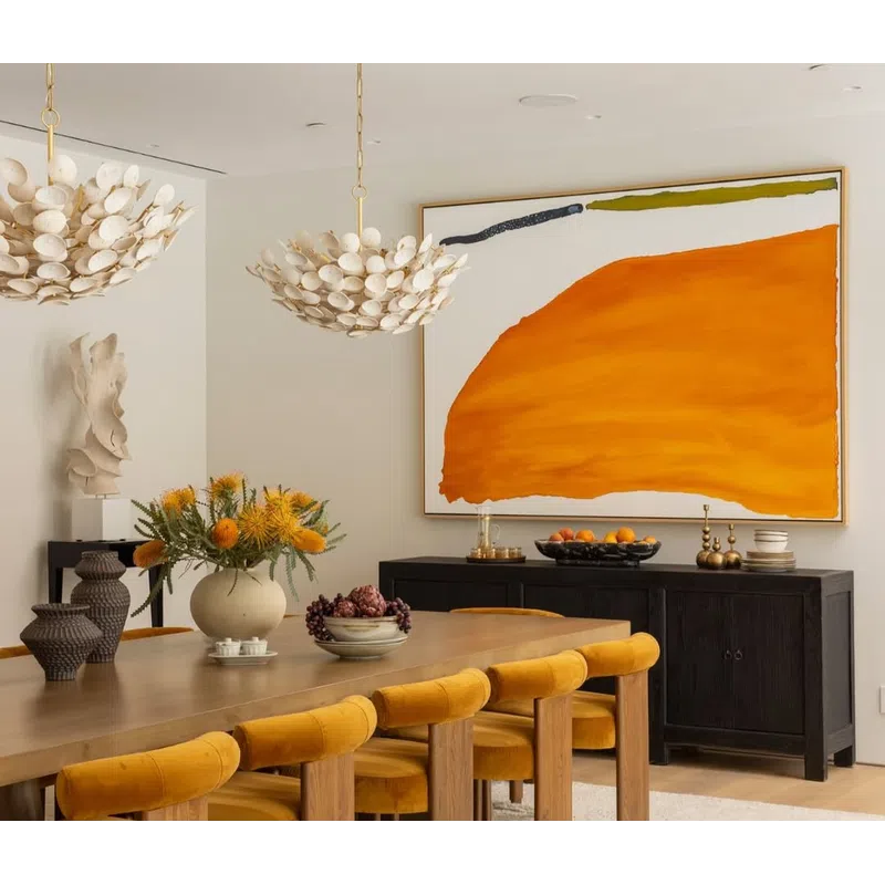 Modern dining room with mustard velvet chairs, wooden table, abstract orange wall art, and decorative lighting.