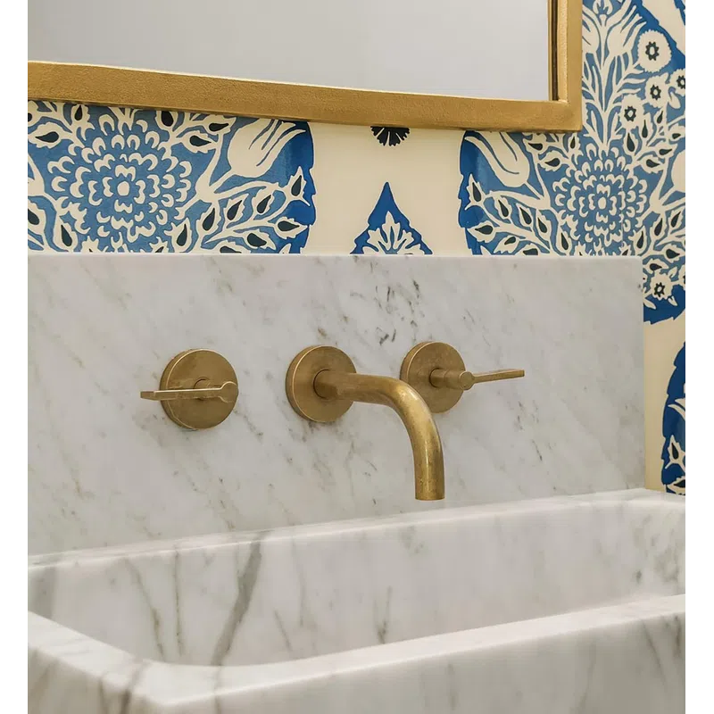 Marble sink with gold faucet set against blue and white floral wallpaper and a gold-framed mirror.