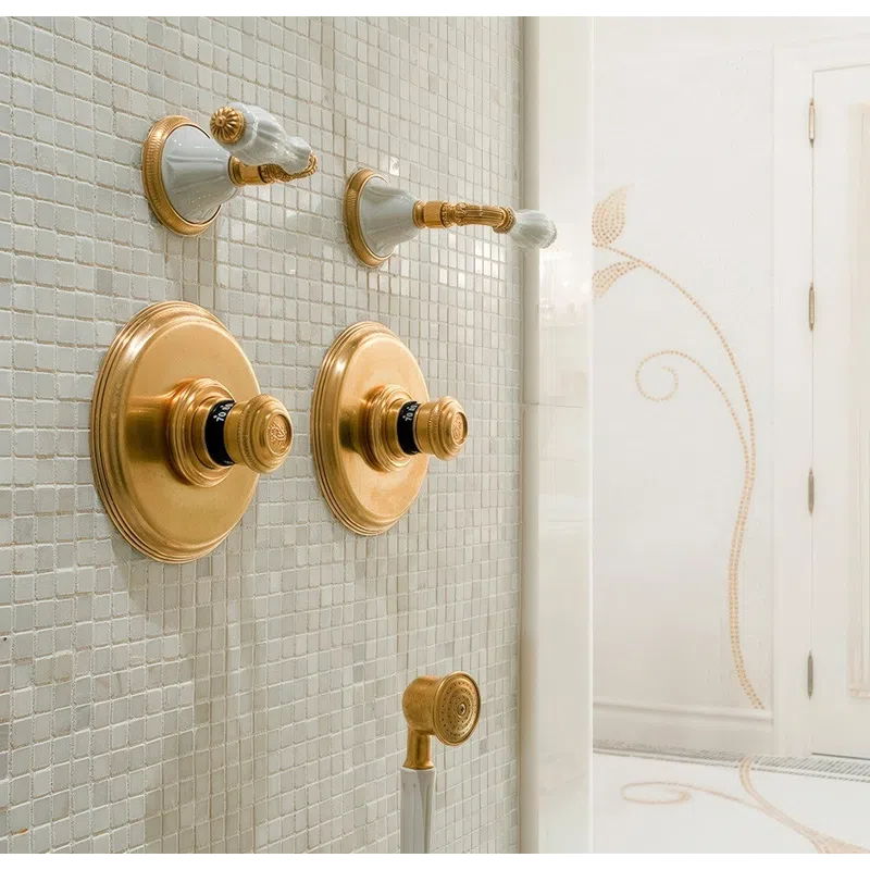 Elegant gold and white bathroom shower controls and hand shower against a tiled wall with decorative floor and door.