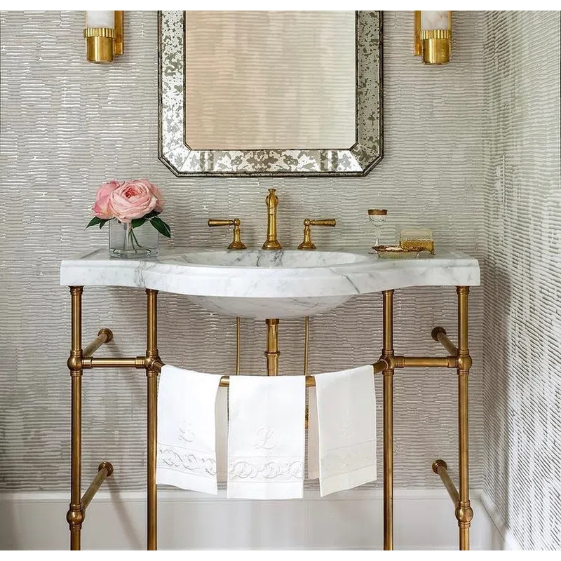 Elegant bathroom vanity with marble sink, gold fixtures, floral arrangement, mirror, and textured wall lighting.