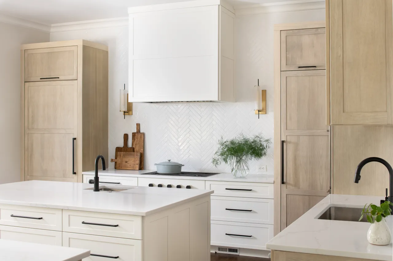 Modern white kitchen with wooden cabinets, marble countertops, dual black faucets, and green plant decoration.