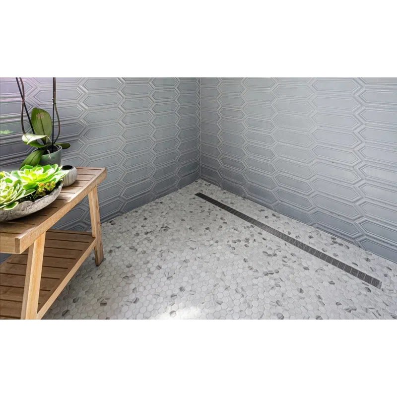 Modern shower with gray hexagonal wall tiles, pebble-style floor, wooden bench, and purple orchids near frosted window.