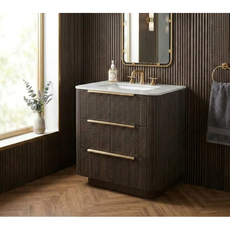 Modern bathroom vanity with ribbed dark wood cabinetry, brass hardware, white countertop, mirror, and natural light window.
