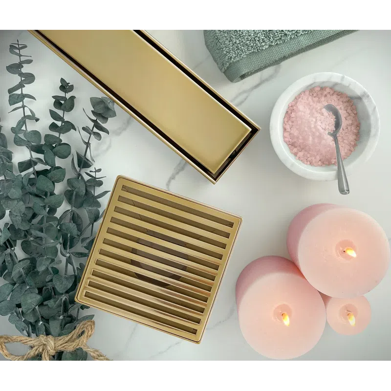 Gold shower drain grate with pink candles, eucalyptus branches, green towel, and bowl of pink bath salts on marble.