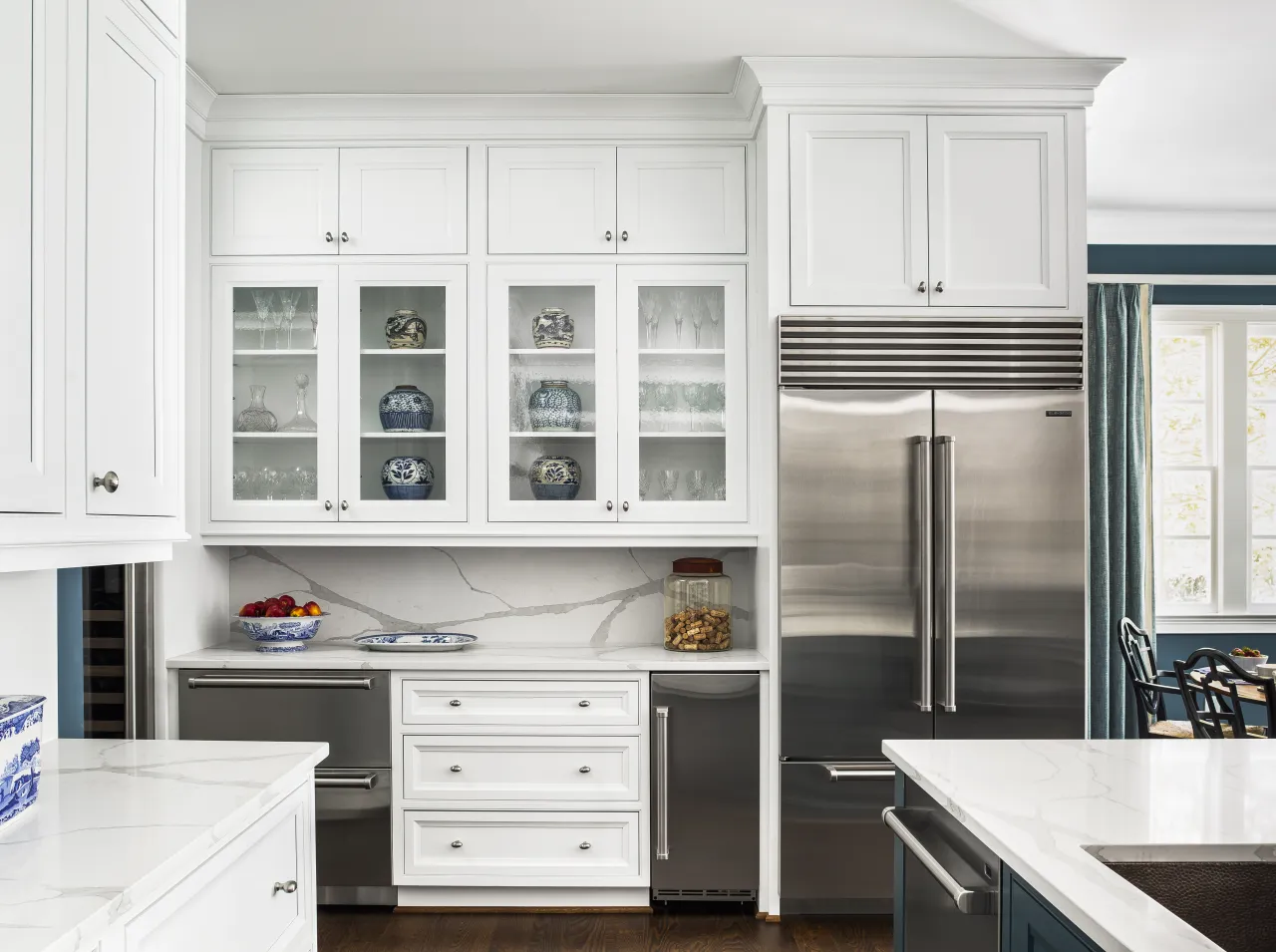 Modern white kitchen with glass-front cabinets, stainless steel appliances, marble countertops, and blue decorative vases.