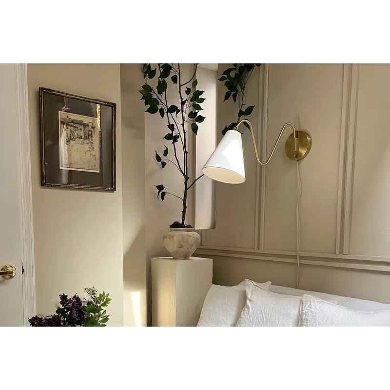 Cozy corner with white couch, three pillows, wall lamp, potted plant, dark wooden table, and framed artwork on beige walls.