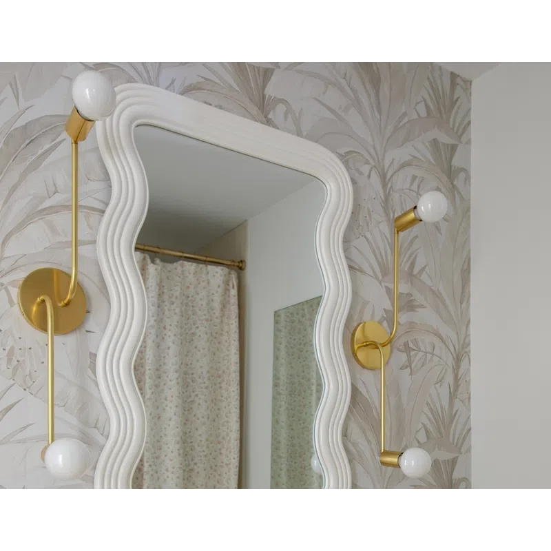 Elegant bathroom with wavy white mirror, gold fixtures, floral wallpaper, and beige marble countertop with sink.