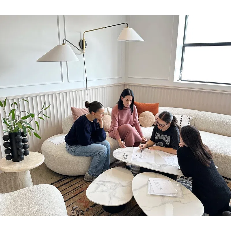 Four women collaborating over architectural plans in a modern, bright living room with contemporary furniture.