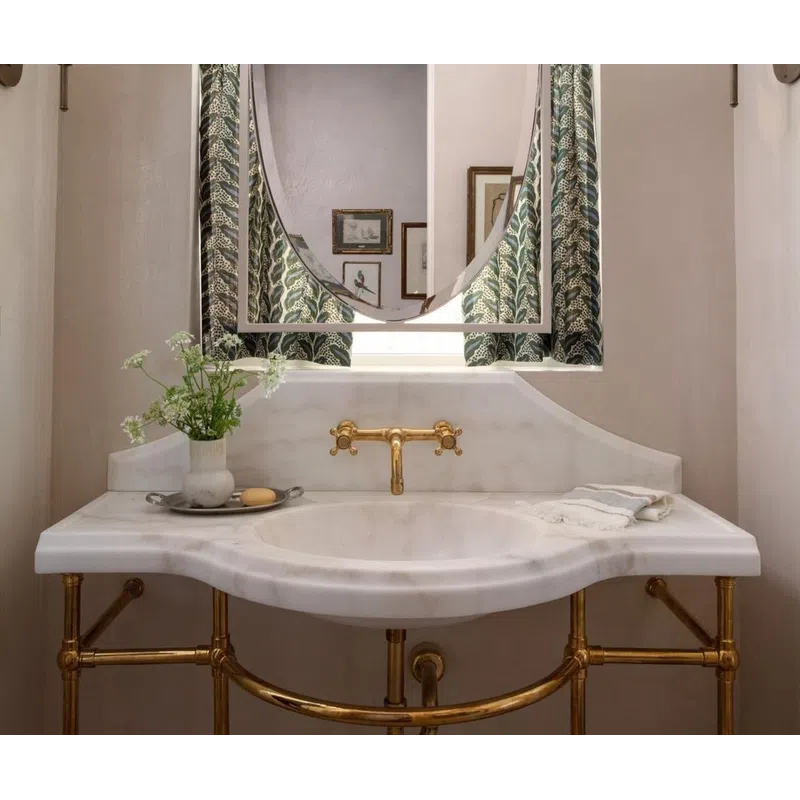 Elegant bathroom sink with gold fixtures, oval mirror, patterned curtains, wall sconces, and decorative vase.