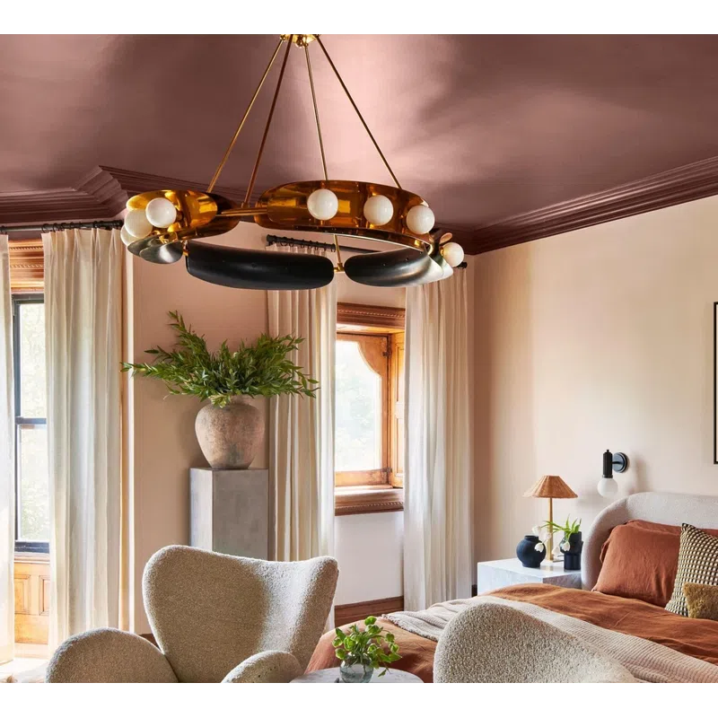 Cozy bedroom with beige armchairs, rust-colored bedding, large windows, and modern chandelier lighting.