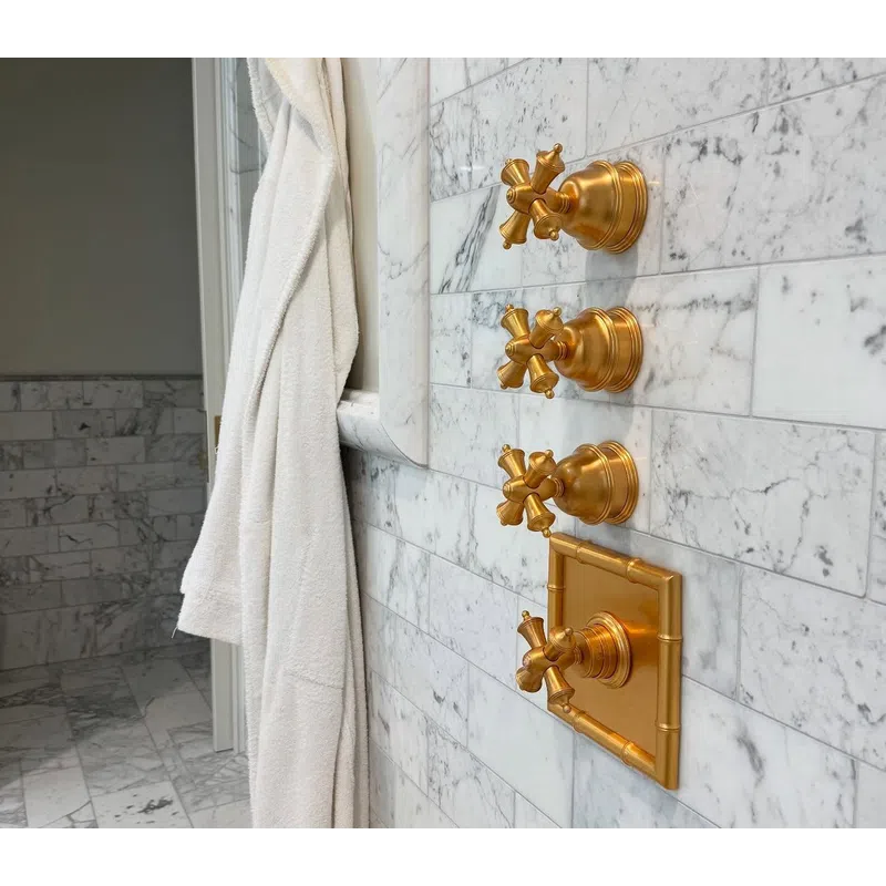 White bathrobe hanging next to four gold vintage shower controls on a marble tiled wall in a bathroom.
