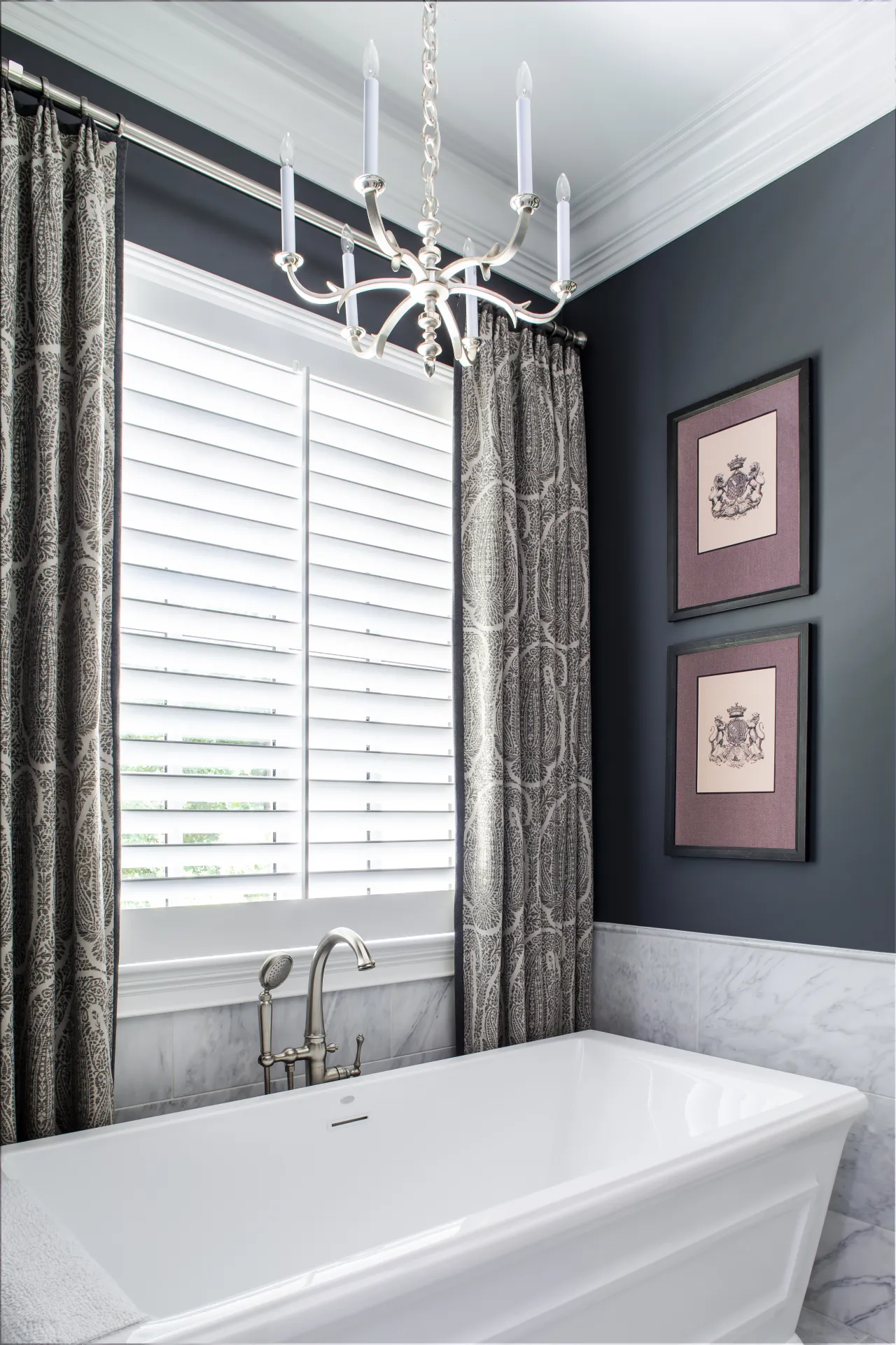 Modern bathroom with white bathtub, silver faucet, patterned curtains, chandelier, and framed wall art.