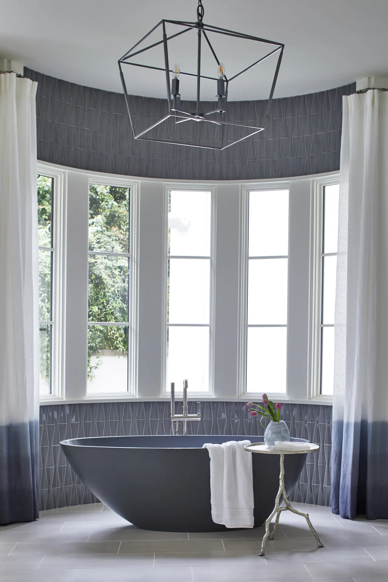 Modern bathroom with a black freestanding bathtub, large windows, white curtains, and geometric tile walls.