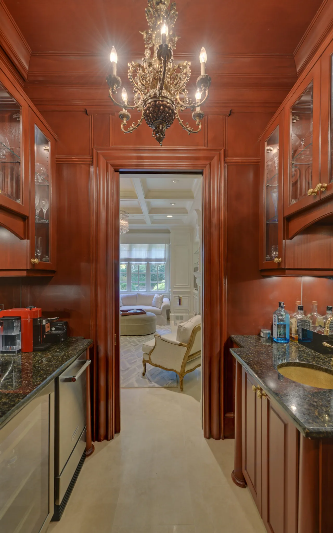 Luxurious wet bar with dark wood cabinets, granite countertops, chandelier, and view into elegant living room.