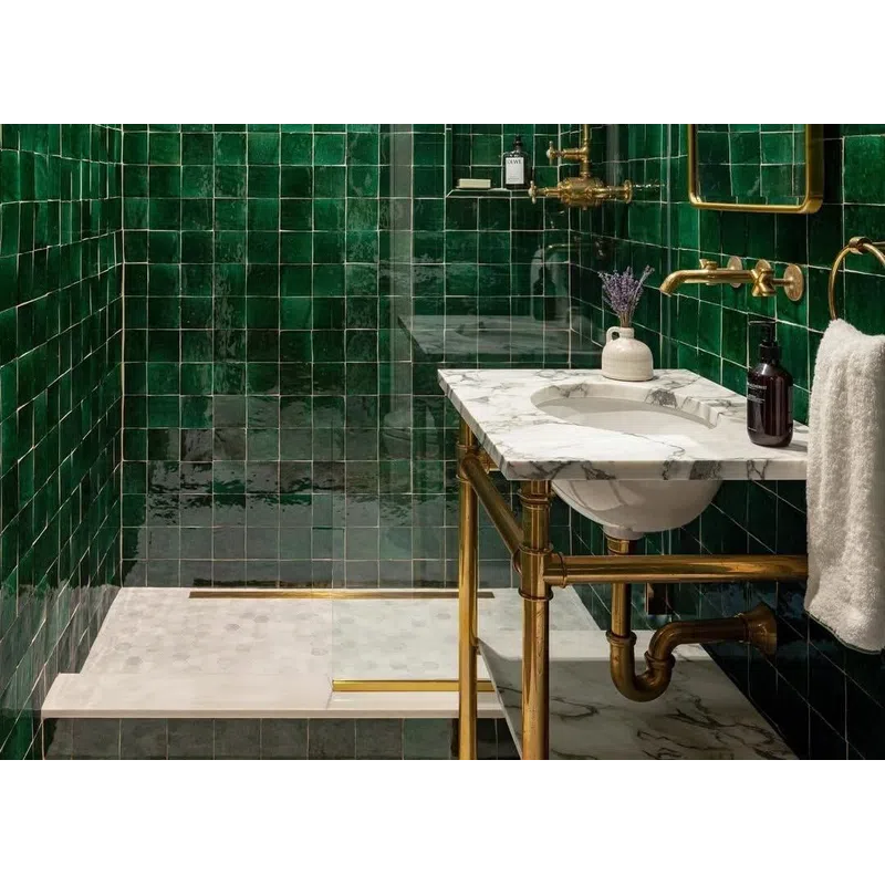 Modern bathroom with glossy green tiles, brass fixtures, marble countertop, and a glass-enclosed shower area.