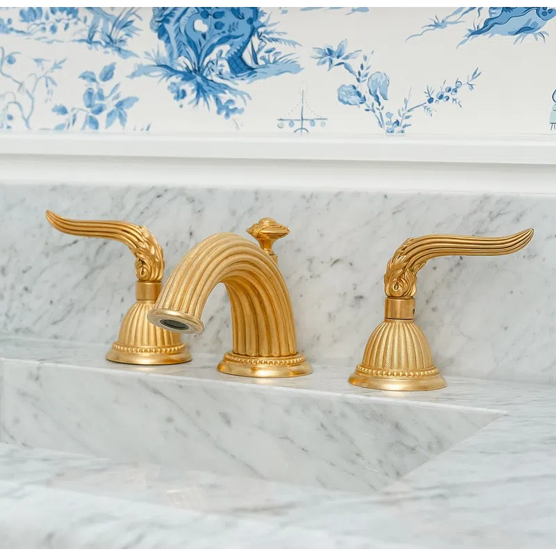 Elegant gold faucet with winged handles on a marble sink under a gold-framed mirror and blue floral wallpaper.