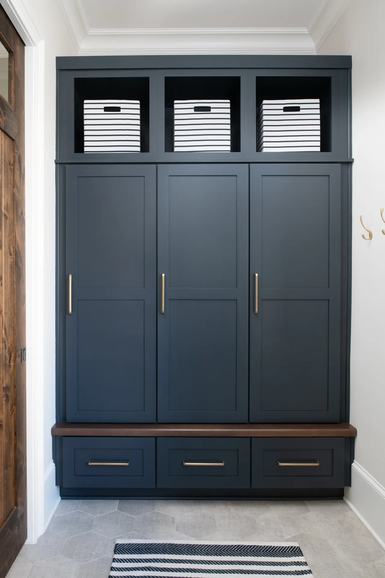 Dark blue built-in storage cabinet with wooden bench, striped storage bins, and brass handles in bright entryway