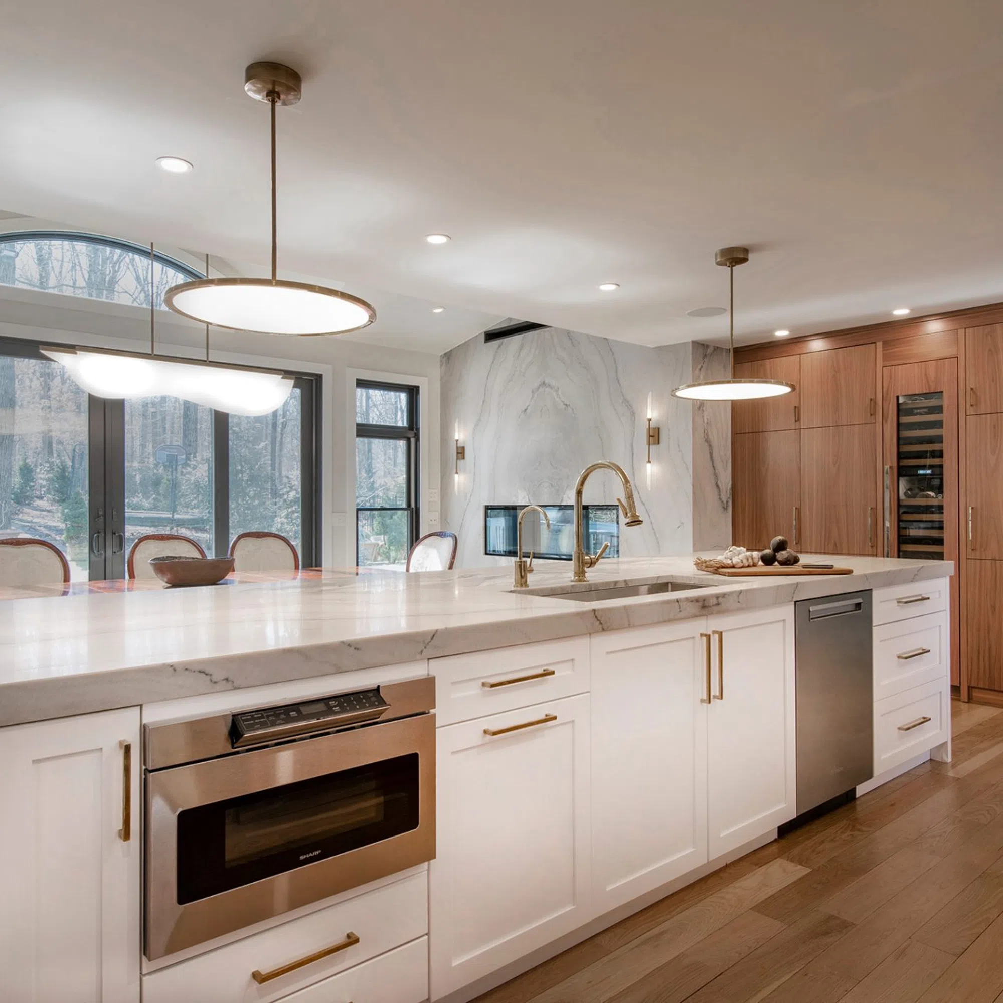 Modern kitchen with white marble island, brass fixtures, wood cabinets, stainless appliances, and large windows.