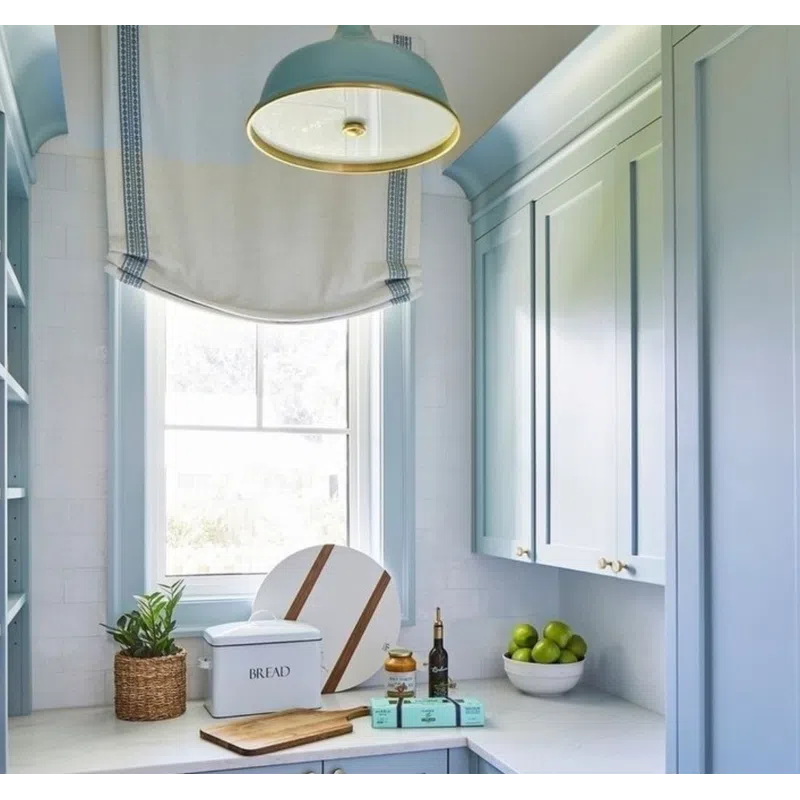 Bright kitchen with blue cabinets, white countertops, window with Roman shade, and countertop decor including bread box and fruit bowl.