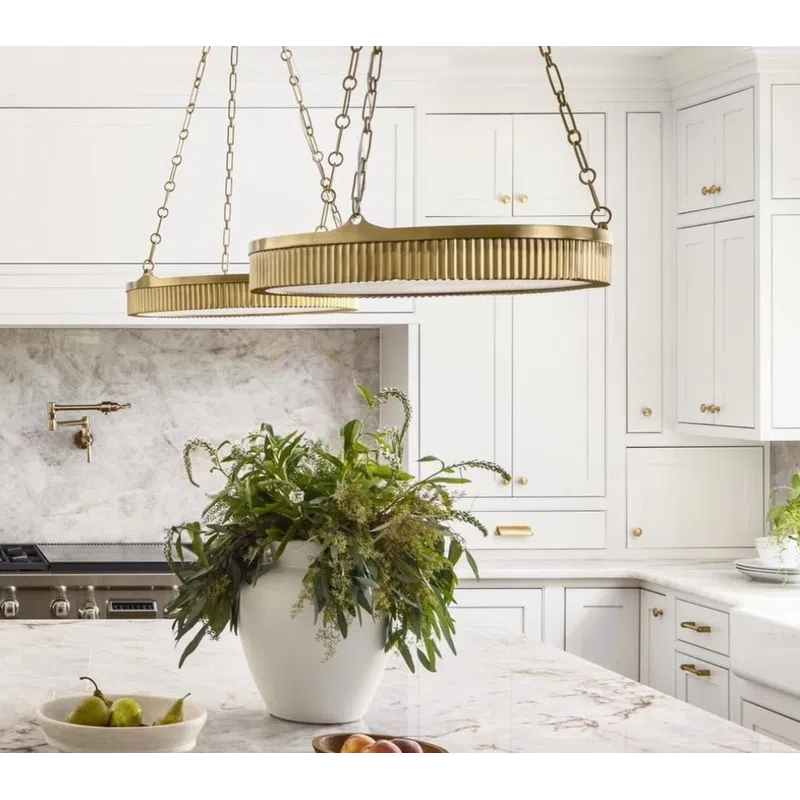 White kitchen with marble countertops, brass pendant lights, green plant centerpiece, and fruit bowls.
