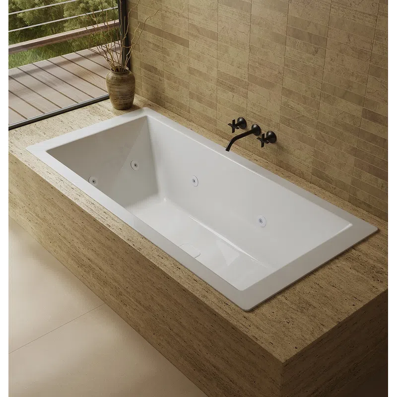 Modern white bathtub with sleek black faucet set in beige stone surround next to a window with potted plant.