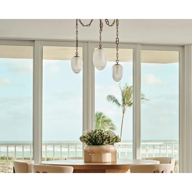 Modern dining area with wooden table, cozy chairs, pendant lights, and ocean view through large windows.