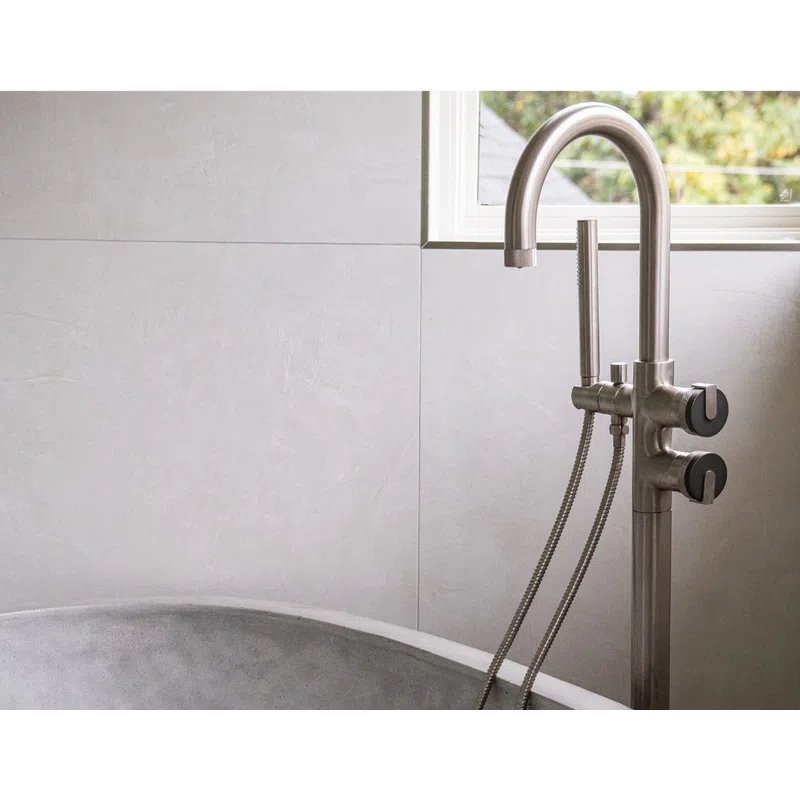 Modern concrete bathtub with stainless steel freestanding faucet and handheld shower against tiled wall.