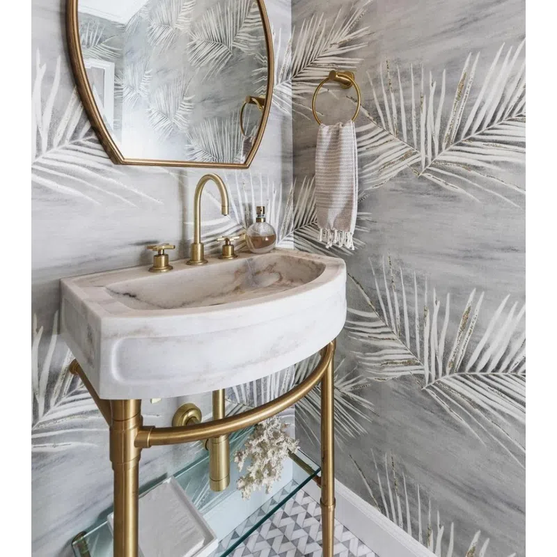 Modern bathroom with marble sink, gold fixtures, leaf-patterned wallpaper, and oval mirror under round light.